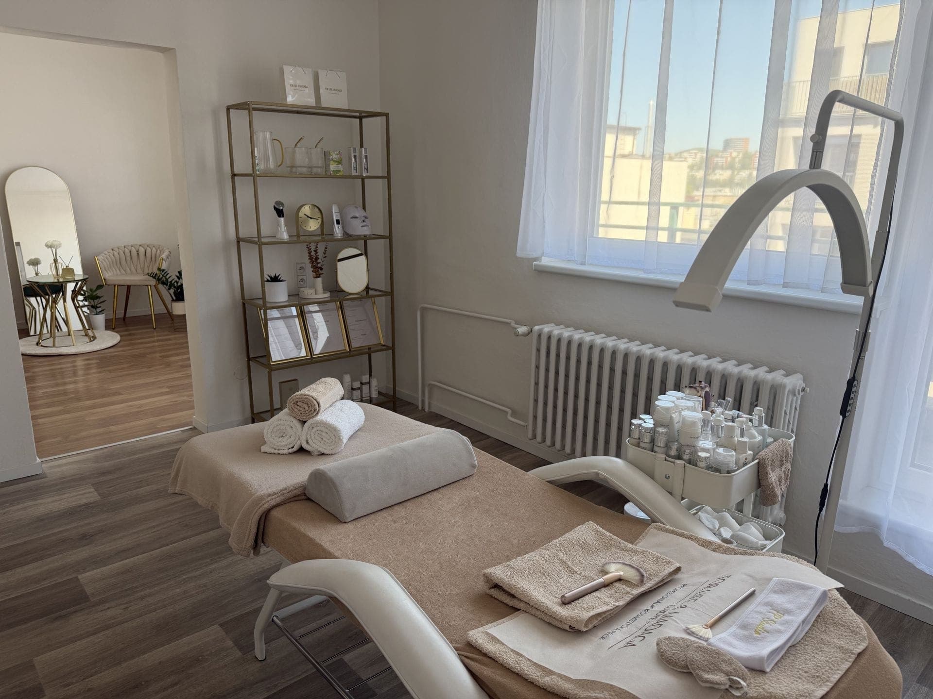 Kosmetické studio s připraveným lehátkem, ručníky, kosmetickými štětci, produkty na pojízdném stolku, profesionální lampou a regálem s dekoracemi.